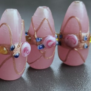 Pode incluir: Três contas de vidro rosa com redemoinhos e detalhes dourados. Cada conta tem um design central de um redemoinho branco e rosa, rodeado por detalhes azuis e amarelos. As contas têm formato oval.