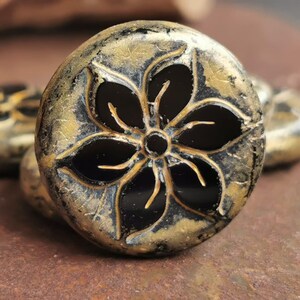 May include: Close-up of a decorative, round bead with a black flower design. The bead has a metallic gold and black finish, with a textured surface. Other similar beads are visible in the background.
