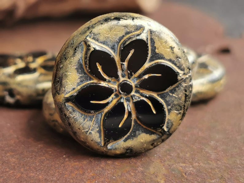 May include: Close-up of a decorative, round bead with a black flower design. The bead has a metallic gold and black finish, with a textured surface. Other similar beads are visible in the background.