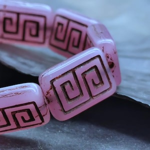 Puede incluir: Primer plano de una pulsera con cuentas rectangulares rosas con un diseño de llave griega marrón oscuro. Las cuentas están ensartadas, mostrando un patrón repetitivo. La pulsera está sobre un fondo borroso.