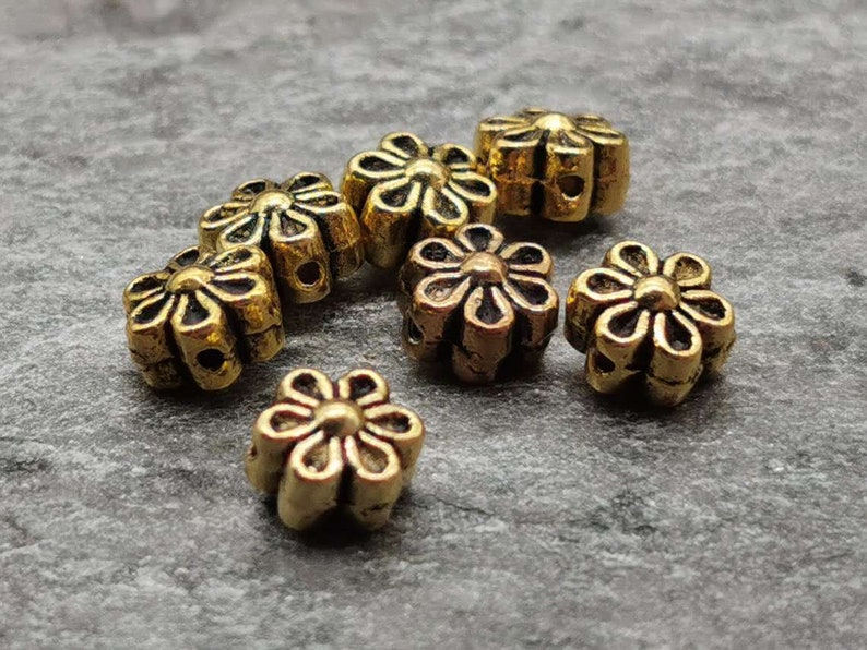 May include: A group of seven gold-colored metal flower-shaped beads. The beads have a raised design and are perfect for jewelry making or crafting.