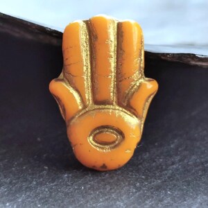 以下が含まれることがあります： 金色のアクセントが付いたオレンジ色のハムサハンドチャームのクローズアップ。保護の象徴であるこのチャームは、手形のペンダントで、ジュエリーやスピリチュアルな実践でよく使用されます。