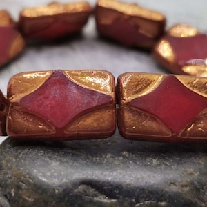 Könnte beinhalten: Nahaufnahme eines Armbands aus roten und goldenen Glasperlen. Die Perlen sind rechteckig mit einer Diamantform in der Mitte. Das Armband liegt auf einer grauen Steinoberfläche.