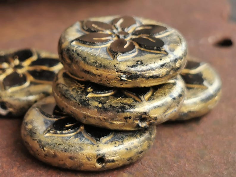 May include: Close-up of several gold and black flower-shaped beads. The beads are stacked, showcasing their detailed floral design. The beads have a metallic finish, adding a touch of elegance. These beads are ideal for jewelry making.