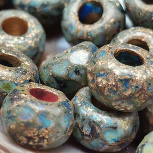 Könnte beinhalten: Nahaufnahme einer Sammlung handgefertigter Perlen. Die Perlen sind eine Mischung aus Türkis, Blau und Gold mit einer strukturierten, unregelmäßigen Oberfläche. Jede Perle hat ein großes zentrales Loch. Ideal für Schmuckherstellung.