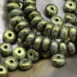 Könnte beinhalten: Nahaufnahme von olivgrünen und dunkelgrauen Perlen mit gesprenkeltem Muster. Die runden Perlen haben ein Loch in der Mitte und eignen sich für Schmuckherstellung oder Bastelarbeiten.