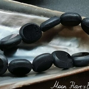 Peut inclure: Un collier de perles ovales noires, en pierre polie, disposées en cercle sur une surface texturée et irisée.