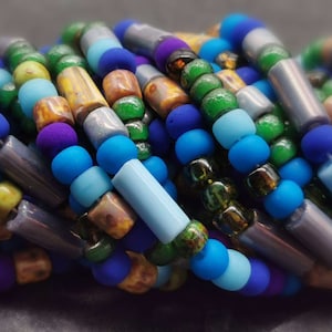 Könnte beinhalten: Nahaufnahme einer Sammlung farbenfroher Perlen in verschiedenen Formen und Größen. Die Perlen sind in Blau-, Grün-, Lila-, Gelb- und Brauntönen gehalten. Einige Perlen sind rund, andere zylindrisch. Die Perlen sind aufgefädelt und bilden eine strukturierte und lebendige Darstellung.