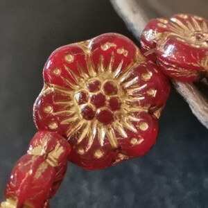 Könnte beinhalten: Nahaufnahme einer Kette aus roten, blütenförmigen Perlen mit goldenen Akzenten. Die Perlen haben ein detailliertes Blumendesign mit einem zentralen Cluster und strahlenförmigen Blütenblättern. Die Perlen sind aufgefädelt, was auf eine Halskette oder ein Armband hindeutet.