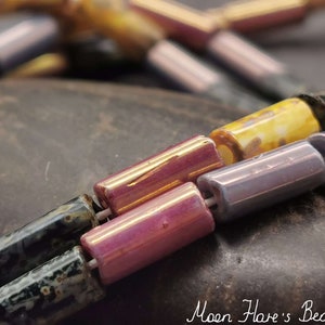 Könnte beinhalten: Nahaufnahme einer Reihe bunter Glasperlen. Die Perlen sind in den Farben Lila, Rosa, Gelb und Schwarz. Die Perlen sind in einem Muster angeordnet und liegen auf einer dunkelgrauen Oberfläche. Der Text "Moon Hare's Beads" ist in der unteren rechten Ecke des Bildes sichtbar.