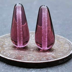 以下が含まれることがあります： ピンクと白の渦巻き模様の入った、涙滴型のガラスビーズが2つ。ビーズの長さは約2.5cmで、銀貨の上に置かれています。