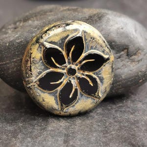 May include: A round, decorative stone with a black flower design. The flower has five petals and a central circle. The stone has a gold and black color scheme and is resting on a larger gray stone.