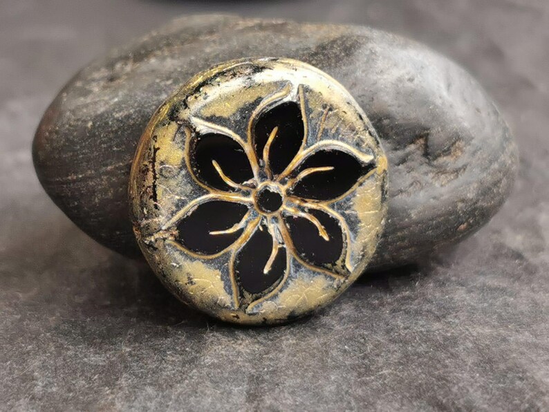 May include: A round, decorative stone with a black flower design. The flower has five petals and a central circle. The stone has a gold and black color scheme and is resting on a larger gray stone.