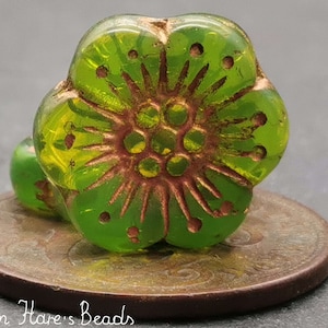 Può includere: Un perlino di vetro verde a forma di fiore con un disegno color rame al centro. Il perlino ha un motivo a stella attorno al disegno centrale.