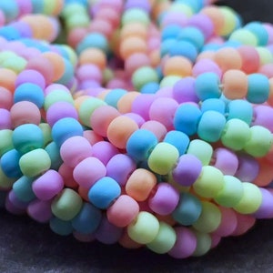 Puede incluir: Primer plano de una hebra de cuentas de vidrio de color pastel. Las cuentas son redondas y tienen un acabado mate. Los colores incluyen azul claro, verde claro, rosa claro y naranja claro.