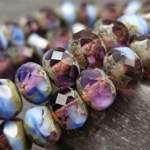 May include: Close-up of faceted glass beads with a purple, blue, and green iridescent finish. The beads have a mottled, marbled effect.