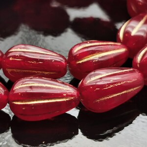 May include: Close-up of several red glass beads with gold stripes. The beads are teardrop-shaped and have a glossy finish. The gold stripes run horizontally across the beads, adding a decorative element. The beads are likely for jewelry making.