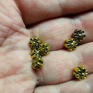 May include: A close-up of a hand holding several gold-colored metal flower-shaped beads. The beads have a slightly raised design and are perfect for jewelry making or crafting.