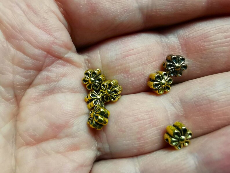 May include: A close-up of a hand holding several gold-colored metal flower-shaped beads. The beads have a slightly raised design and are perfect for jewelry making or crafting.
