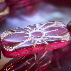 Könnte beinhalten: Nahaufnahme von mehreren rechteckigen, dunkelroten Perlen mit einem floralen Design in einer helleren, metallischen Farbe. Die Perlen haben eine glänzende Oberfläche und sind eng aneinandergereiht, was auf eine Halskette oder ein Armband hindeutet.