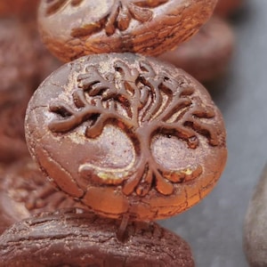 Peut inclure: Gros plan sur une perle en verre marron et or avec un motif d'arbre de vie. La perle est texturée et a un aspect rustique.