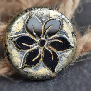 May include: A round, decorative button with a black floral design. The button has a metallic gold and silver finish with a distressed look. The button is likely used for sewing or crafts.