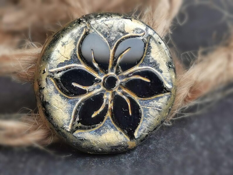 May include: A round, decorative button with a black floral design. The button has a metallic gold and silver finish with a distressed look. The button is likely used for sewing or crafts.