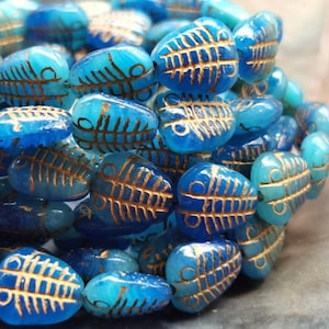 Könnte beinhalten: Nahaufnahme einer Sammlung blauer und türkisfarbener Glasperlen. Jede Perle hat ein fischähnliches Design in Gold. Die Perlen sind oval geformt und scheinen aufgefädelt zu sein, möglicherweise für die Schmuckherstellung oder das Basteln.