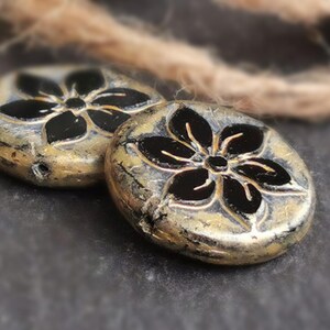 May include: Two round, metallic beads with a flower design. The beads have a gold and silver finish with a black flower in the center. The beads are on a dark gray surface with a brown rope in the background.