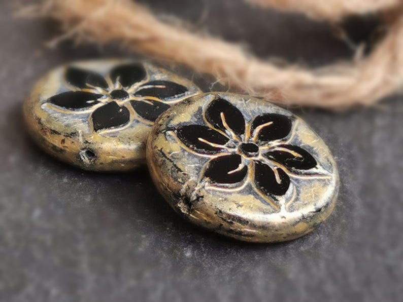 May include: Two round, metallic beads with a flower design. The beads have a gold and silver finish with a black flower in the center. The beads are on a dark gray surface with a brown rope in the background.