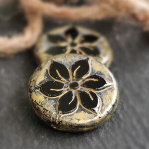 May include: Close-up of two round, antique-style beads. Each bead features a black floral design on a gold-colored base. The beads have a distressed, textured appearance, suggesting an aged or vintage aesthetic. A neutral-colored string is in the background.