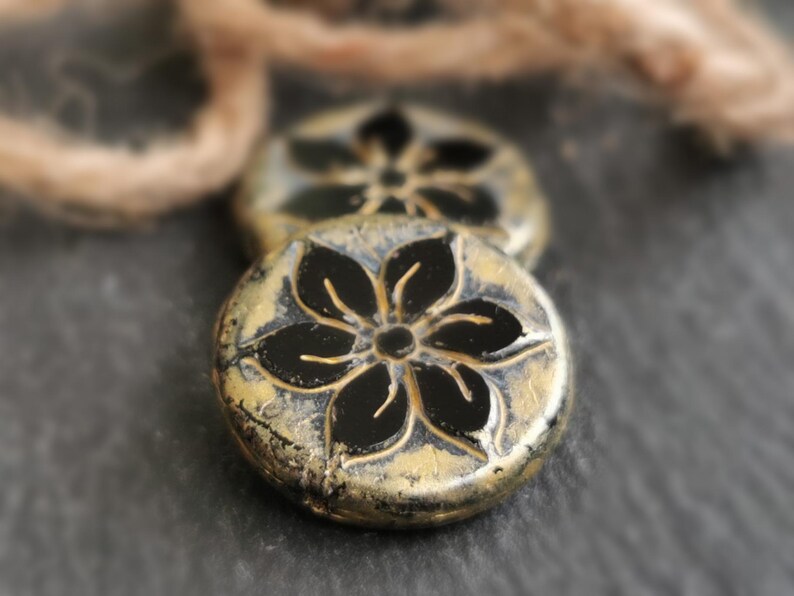 May include: Close-up of two round, antique-style beads. Each bead features a black floral design on a gold-colored base. The beads have a distressed, textured appearance, suggesting an aged or vintage aesthetic. A neutral-colored string is in the background.