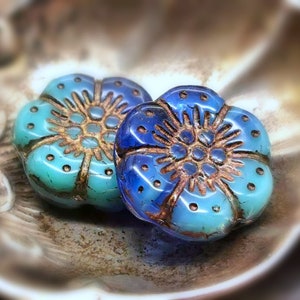 Può includere: Due perline a forma di fiore nelle tonalità del blu e del turchese con un centro color bronzo. Le perline hanno una superficie strutturata e sono esposte su un piatto decorativo in metallo. Il design floreale presenta petali dettagliati e un motivo centrale.