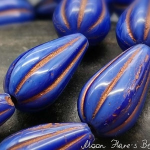 May include: A close-up of blue glass beads with copper-colored stripes. The beads are shaped like teardrops and have a glossy finish.