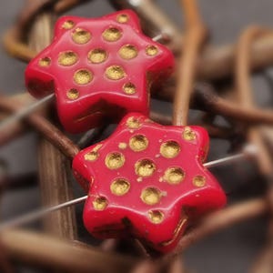 Könnte beinhalten: Zwei rote sternförmige Perlen mit goldenen Punkten. Die Perlen sind aus Glas und haben eine glänzende Oberfläche.