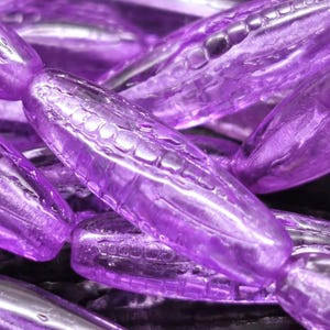 May include: Close-up of several translucent purple beads. The beads are oblong with a textured surface and are strung together. The lighting creates highlights and shadows, enhancing the depth and detail of the beads. The beads are a vibrant shade of purple.
