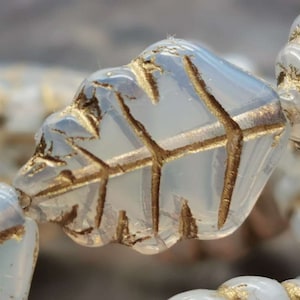 Könnte beinhalten: Nahaufnahme einer weißen Glasperle mit einem goldenen Blattdesign. Die Perle ist Teil einer Perlenkette.