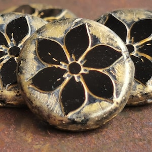 May include: Close-up of several round, decorative beads. Each bead features a black flower design on a gold-colored background. The beads have a textured, metallic appearance, suggesting they are made of metal or have a metallic finish.