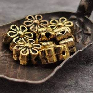 May include: A group of gold-colored metal flower-shaped beads. The beads are arranged in a pile on a dark brown surface.