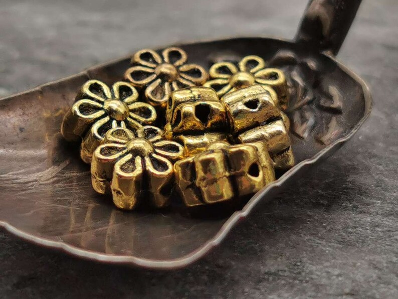 May include: A group of gold-colored metal flower-shaped beads. The beads are arranged in a pile on a dark brown surface.