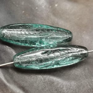 May include: Two teal-colored glass beads with an elongated oval shape and a textured, patterned interior. The beads are strung on a thin, silver-colored wire, suitable for jewelry making.