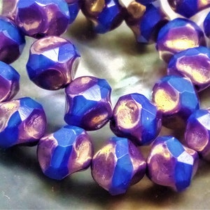 Può includere: Una collana di perline di vetro sfaccettate iridescenti di colore viola e blu. Le perline hanno un effetto metallico e misurano circa 10 mm di diametro.