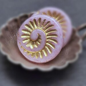 Könnte beinhalten: Zwei rosa Glasknöpfe mit einem goldenen Wirbelmuster. Die Knöpfe liegen auf einer weißen Oberfläche.