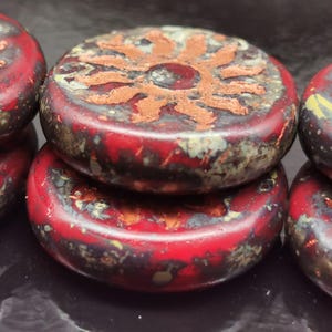 Peut inclure: Gros plan sur plusieurs perles en céramique rouges et marron. Chaque perle a une forme plate et circulaire avec un motif en relief en forme de soleil de couleur marron clair. Les perles ont un aspect vieilli.