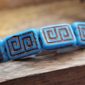 May include: Close-up of a bracelet featuring rectangular turquoise beads with a brown Greek key design. The beads are strung together, showcasing a classic and elegant style. The bracelet is set against a wooden surface.