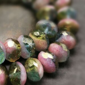 Puede incluir: Primer plano de una hebra de cuentas de vidrio iridiscente. Las cuentas son facetadas y tienen una combinación de colores rosa, verde y dorado.