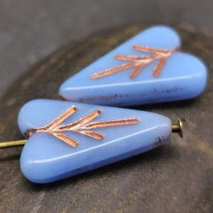 May include: Two light blue glass heart-shaped beads with copper-colored arrow designs. The beads have a hole at the top for stringing.