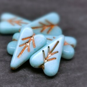Può includere: Un gruppo di sei perline di vetro di colore blu chiaro a forma di cuore con un motivo a freccia di colore rame.