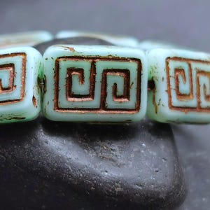 Puede incluir: Primer plano de un hilo de cuentas cuadradas. Cada cuenta es de color verde pálido con un diseño de llave griega color cobre. Las cuentas descansan sobre una piedra gris oscuro, creando un contraste de color y textura.