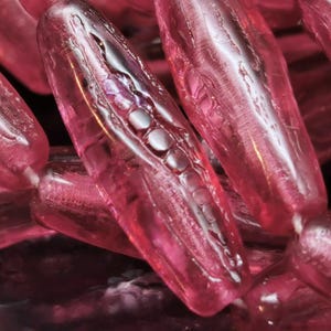 May include: Close-up of several translucent, oblong, pink beads. Each bead has a series of small, round, white accents. The beads are clustered together, creating a textured and layered effect. The lighting highlights the glossy surface.
